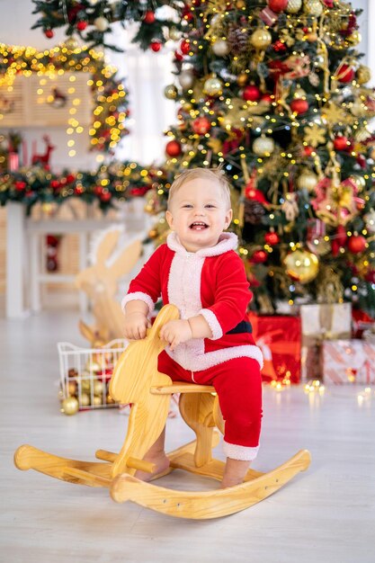 A cute little boy in a Santa costume rides a rocking toy under a festive Christmas tree with gifts in the living room of the house a happy kid celebrates Christmas and New Year at home