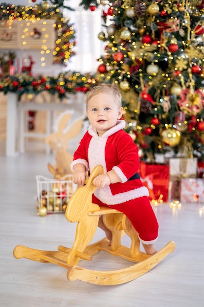 A cute little boy in a Santa costume rides a rocking toy under a festive Christmas tree with gifts in the living room of the house a happy kid celebrates Christmas and New Year at home