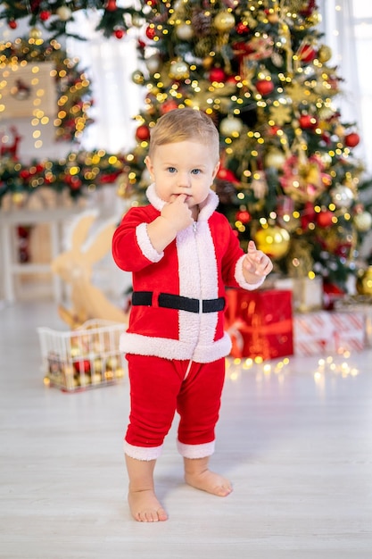 산타 의상을 입은 귀여운 소년이 집 거실에 선물을 들고 축제 크리스마스 트리 아래에 서 있습니다. 행복한 아이는 집에서 크리스마스와 새해를 축하합니다