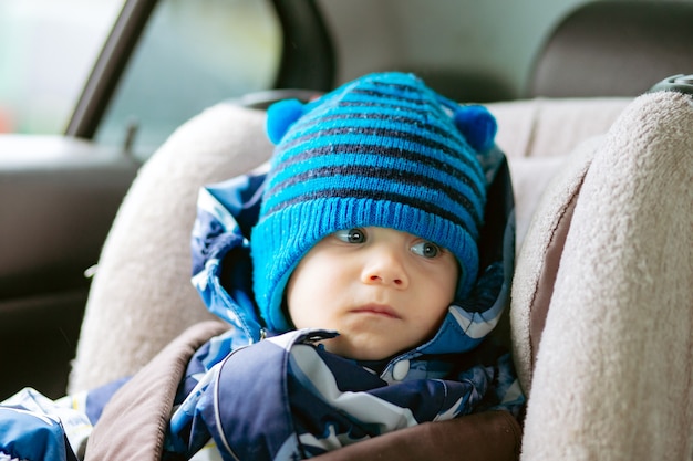 cute little boy sitting in child safety seat in a car wearing winter overall and hat image with sele...