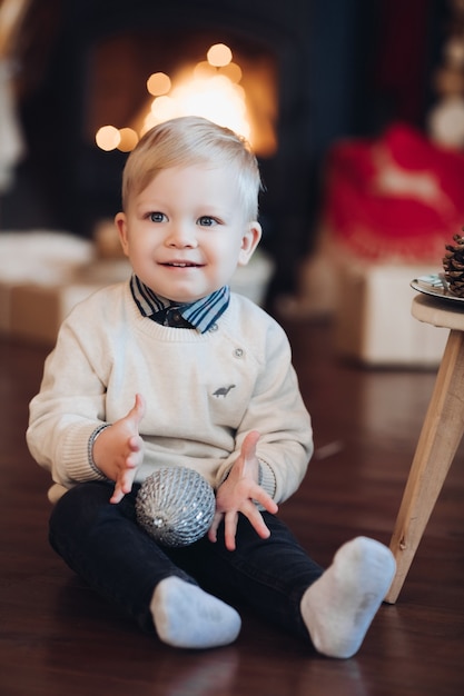 Cute little boy sitting down and holding a sparkling ball ornament with fireplace