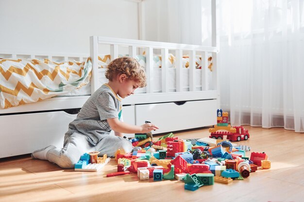 Cute little boy sitting and have fun indoors in the bedroom with plastic construction set