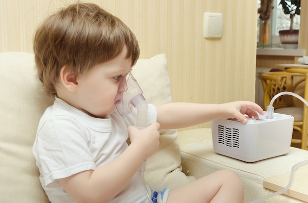 Cute little boy sitting at home on the couch and breathing through a nebulizer inhaler. The boy cures the cough inhaler.
