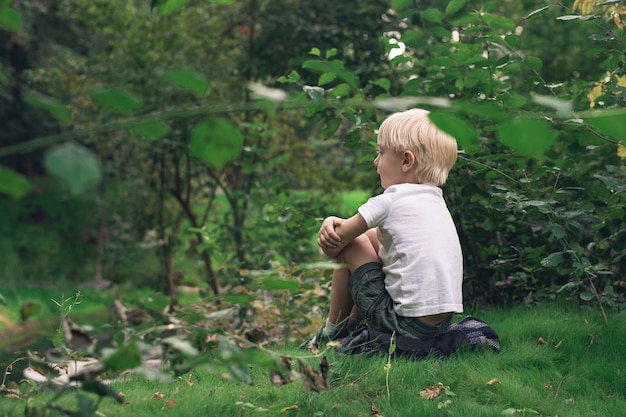 Cute little boy sitting on the lawn in the Park and thinks about life. Children's loneliness concept