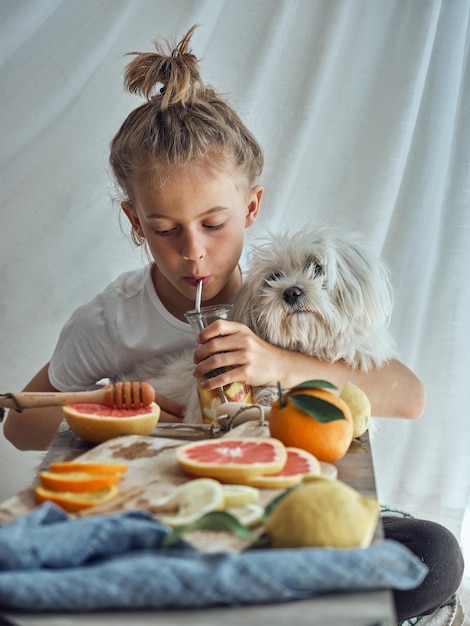 Cute little boy sitting at table and drinking fresh lemonade while spending time with white Yorkshire Terrier