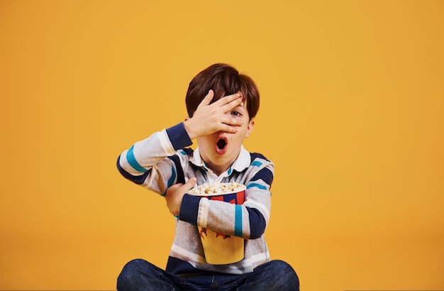 Cute little boy sitting and watching movie and eats popcorn against yellow wall