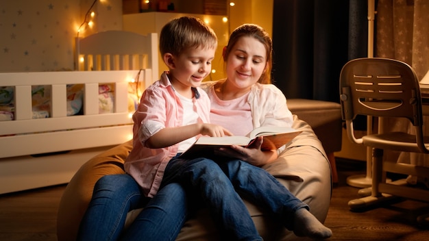 夜に母親と一緒に座って、就寝時の物語の本を読んでいるかわいい男の子。夜に一緒に時間を過ごす子供の教育と家族の概念。