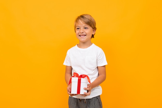 Милый маленький мальчик в футболке и брюках держит коробку с подарком сам изолирован на желтом