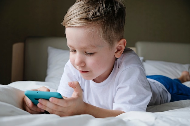 cute little boy toddler playing mobile game, lying on a bed and holding smartphone