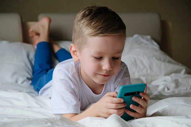 Photo cute little boy toddler playing mobile game, lying on a bed and holding smartphone