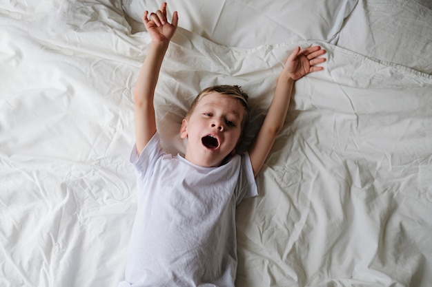 Cute little boy toddler wakes up, yawning and stretching on a bed in a bedroom.