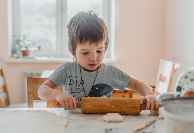 Cute little boy using rolling pin to prepare the dough for cookies in the kitchen