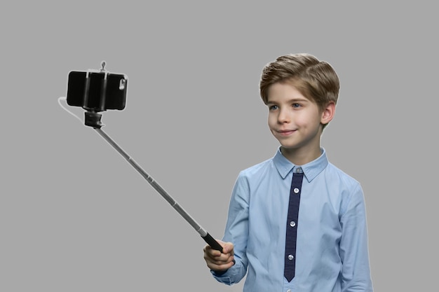 Ragazzino sveglio utilizzando selfie stick. bel bambino con il monopiede di scattare una foto su sfondo grigio. bambini e concetto di tecnologia moderna.