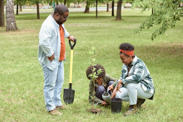 Cute little boy watering small tree planted by his mother in park