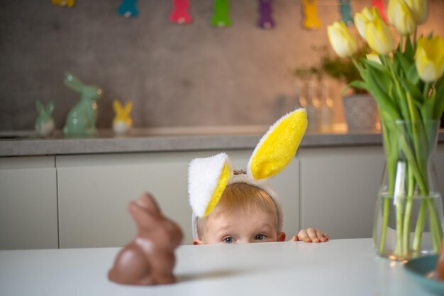 イースターの日にうさぎの耳をかぶったかわいい男の子は、チョコレートのイースターバニーを食べたがっています イースターのためにエッグハントをする子供 自宅でイースターを祝うチャーミングな子供