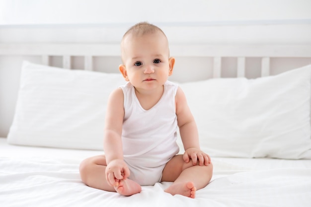 Cute little boy in a white bodysuit sitting on a bed on white bedding in the bedroom portrait of a cute child with brown eyes and blond hair