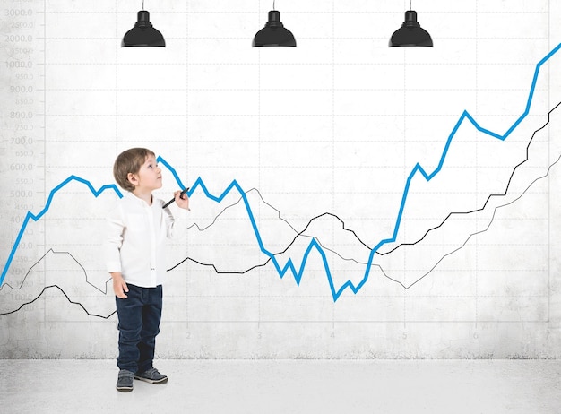 Cute little boy in a white shirt and dark jeans holding a marker and looking upwards. A concrete wall with many graphs drawn on it.