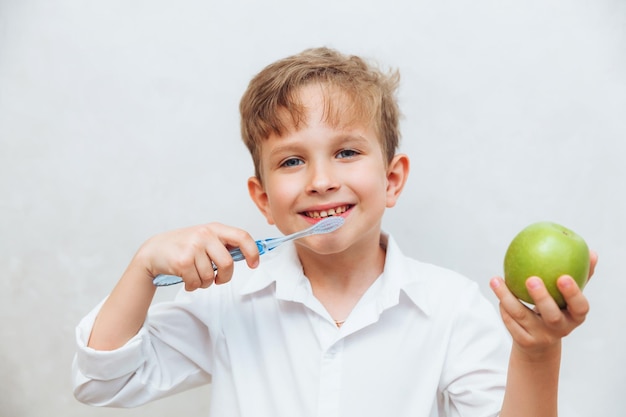 A cute little boy with blond hair and blue eyes is brushing his teeth and holding a big green apple the image is on a white background