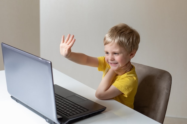 A cute little boy with blond hair sits at a table with a laptop and waves his hand