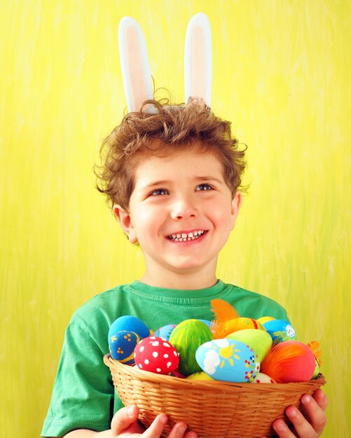 Photo cute little boy with bunny ears on his head and colorful easter eggs in a wicker basket in his hands