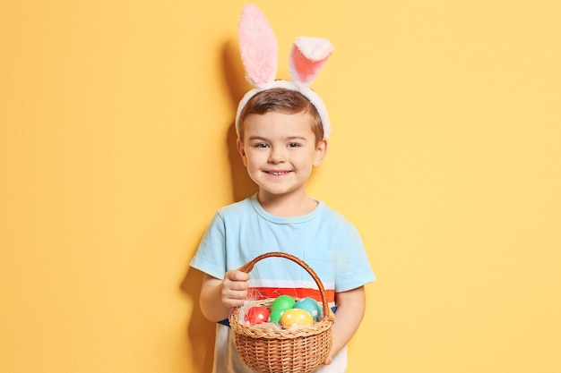 Cute little boy with bunny ears holding basket full of Easter eggs on color