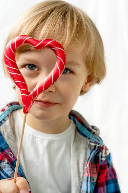 Cute little boy in with candy red lollipops in heart shape, white background. Beautiful kid eat sweets. Valentine's day, love concept.