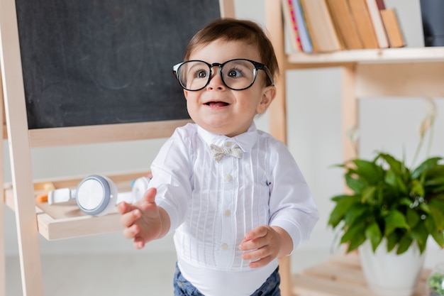 A cute little boy with eye glasses smiling next to the blackboard and books. pre-school education for children