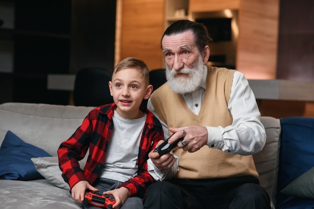 Cute little boy with grandfather sitting on sofa and playing video game with game pad