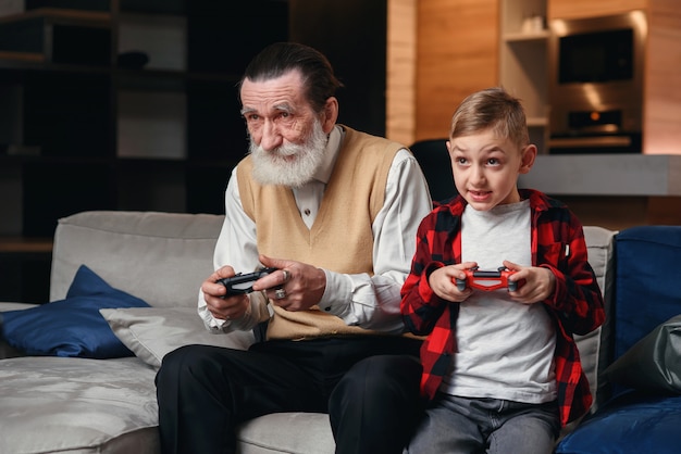 Cute little boy with grandfather sitting on sofa and playing video game with game pad