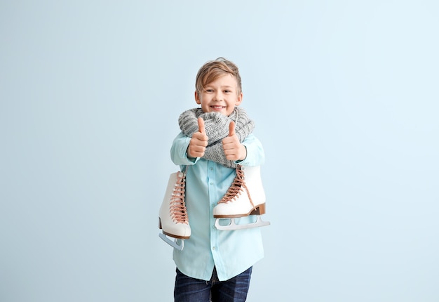 Cute little boy with ice skates showing thumb-up against color