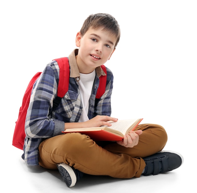 Милый маленький мальчик с красным рюкзаком, читающий книгу, изолированный на белом
