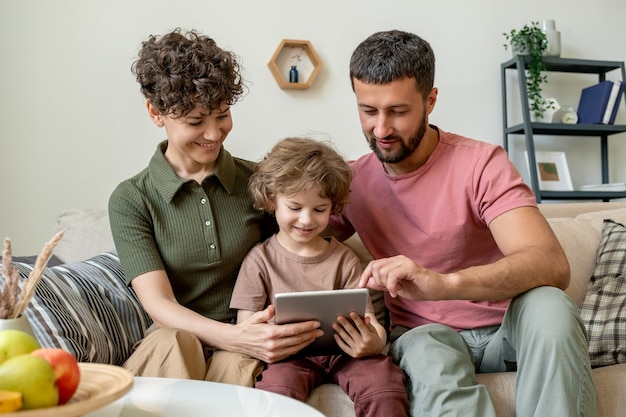 Ragazzino sveglio con il touchpad ei suoi giovani genitori affettuosi che guardano video o film online o cercano cartoni animati curiosi in rete