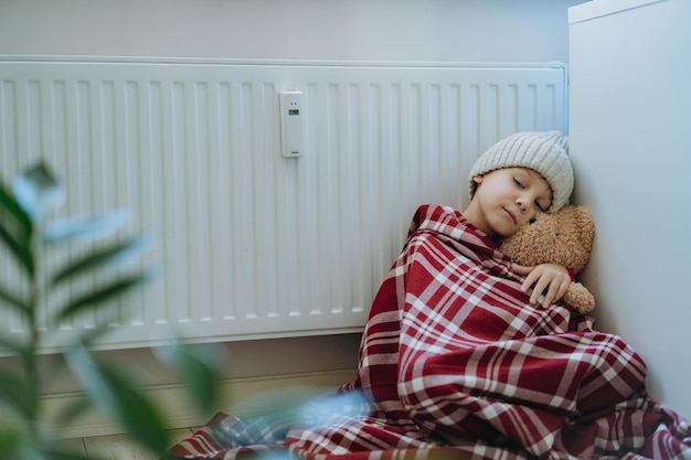 Photo cute little boy wrapped id plaid wearing knit hat sitting by heater hugging teddy bear cold at home