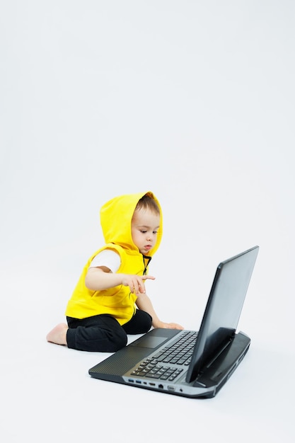A cute little boy in a yellow vest is sitting at a digital laptop on a white background Child and modern portable computer gadget