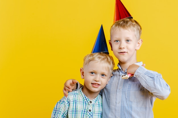 Cute little brothers boys in Birthday hat hug each other on yellow.