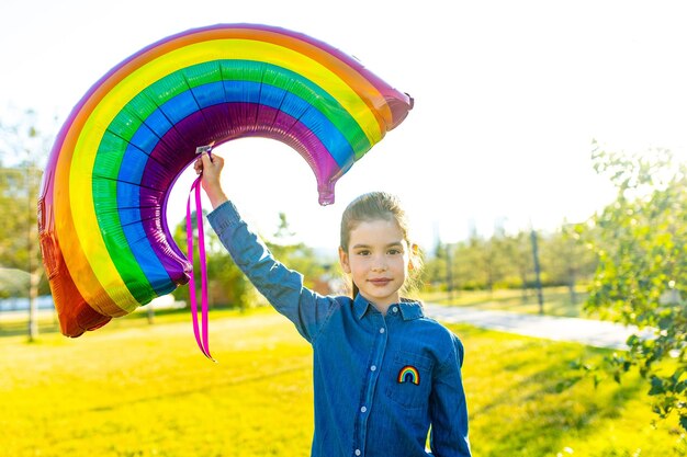 Cute little brunette girl holding an air colorful rainbow balloon in summer park