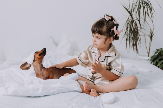 cute little brunette girl at home in bed with a dog playing with baby cosmetics. little lady