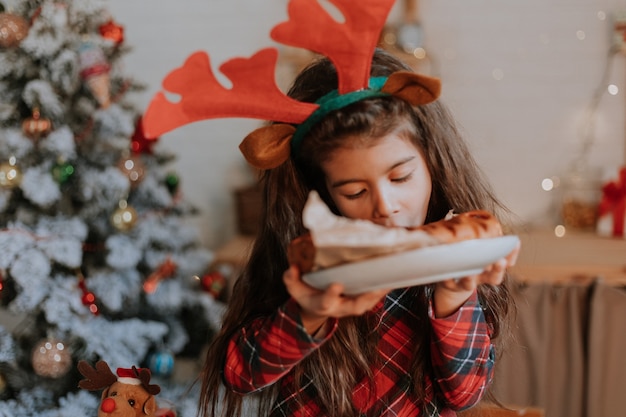 Cute little brunette girl in red pajamas with deer horns on her head is eating a Christmas cake