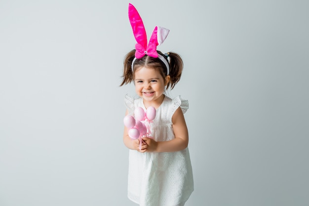 Cute little brunette girl smiling with the ears of a rabbit on her head holding decorative Easter eggs against a light background. concept of Easter, space for text