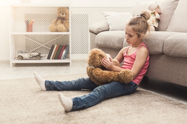 Cute little casual girl embracing teddy bear. Sad kid playing at home, sitting on floor near sofa with her favorite toy, copy space