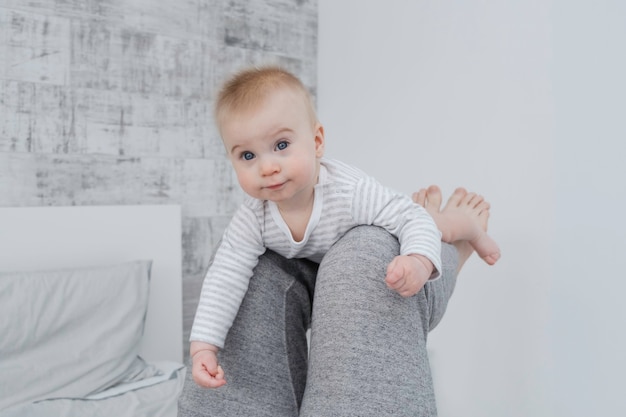 Cute little caucasian baby lying on legs of dad, looking at camera on bed, modern white and gray interior at home.