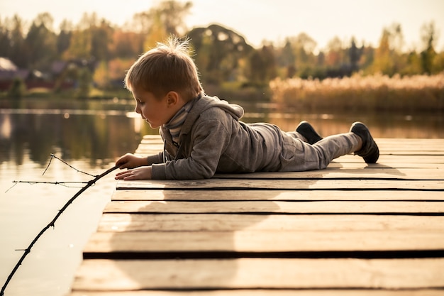 Cute little caucasian boy pretending catching fish in lake laying on wooden pier in the countryside