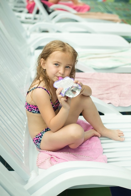 Cute little Caucasian child eats fruit puree in a bag on a chaise longue in a water park