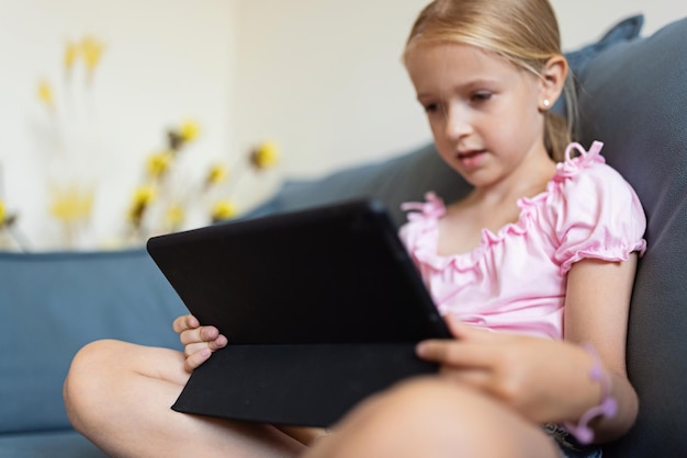 Photo cute little caucasian girl with blonde hair in fashionable clothes sitting at home during coronavirus pandemic quarantine and using tablet                 person