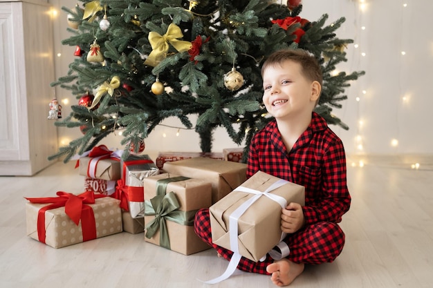 cute little child boy in red pajama with gift boxes sitting under christmas tree at home