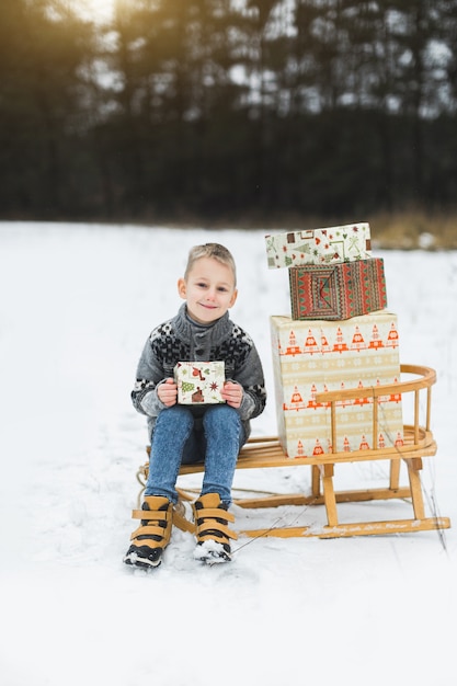 Piccolo bambino sveglio, ragazzo, seduto su una slitta di legno decorata con scatole di regali, tenendo la scatola