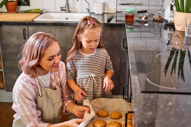 かわいい小さな子供の娘は、お母さんがキッチンオーブンでクッキーを焼くのを手伝って、彼らはそれの準備ができているのを見て立っています