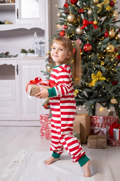 집에서 크리스마스 트리 아래 선물 상자가 있는 크리스마스 잠옷을 입은 귀여운 어린 소녀