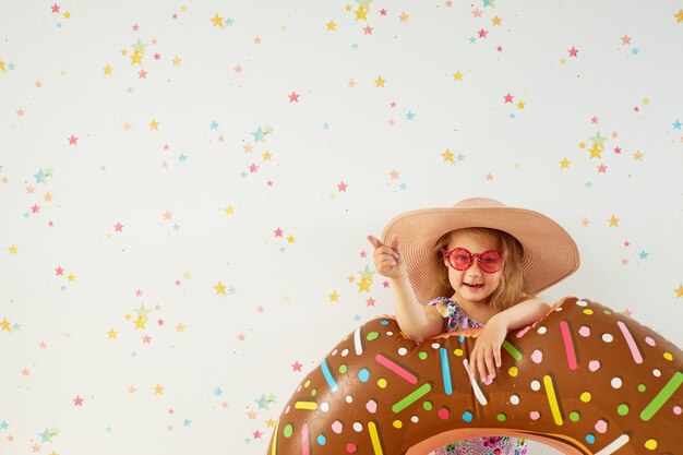 Cute little child girl  in hat with inflatable ring on color wall. Quarantine summer vacation at home