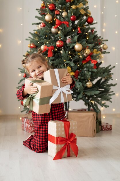 Carina bambina in pigiama rosso con scatole regalo seduta sotto l'albero di natale a casa
